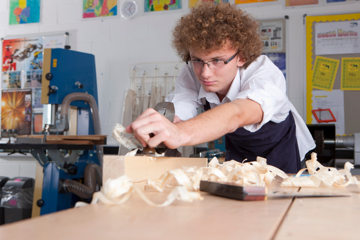 A teenage boy with curly hair wears safety glasses and smirks while using a wood planer to cut wood into the right size in what looks like a high school shop class.