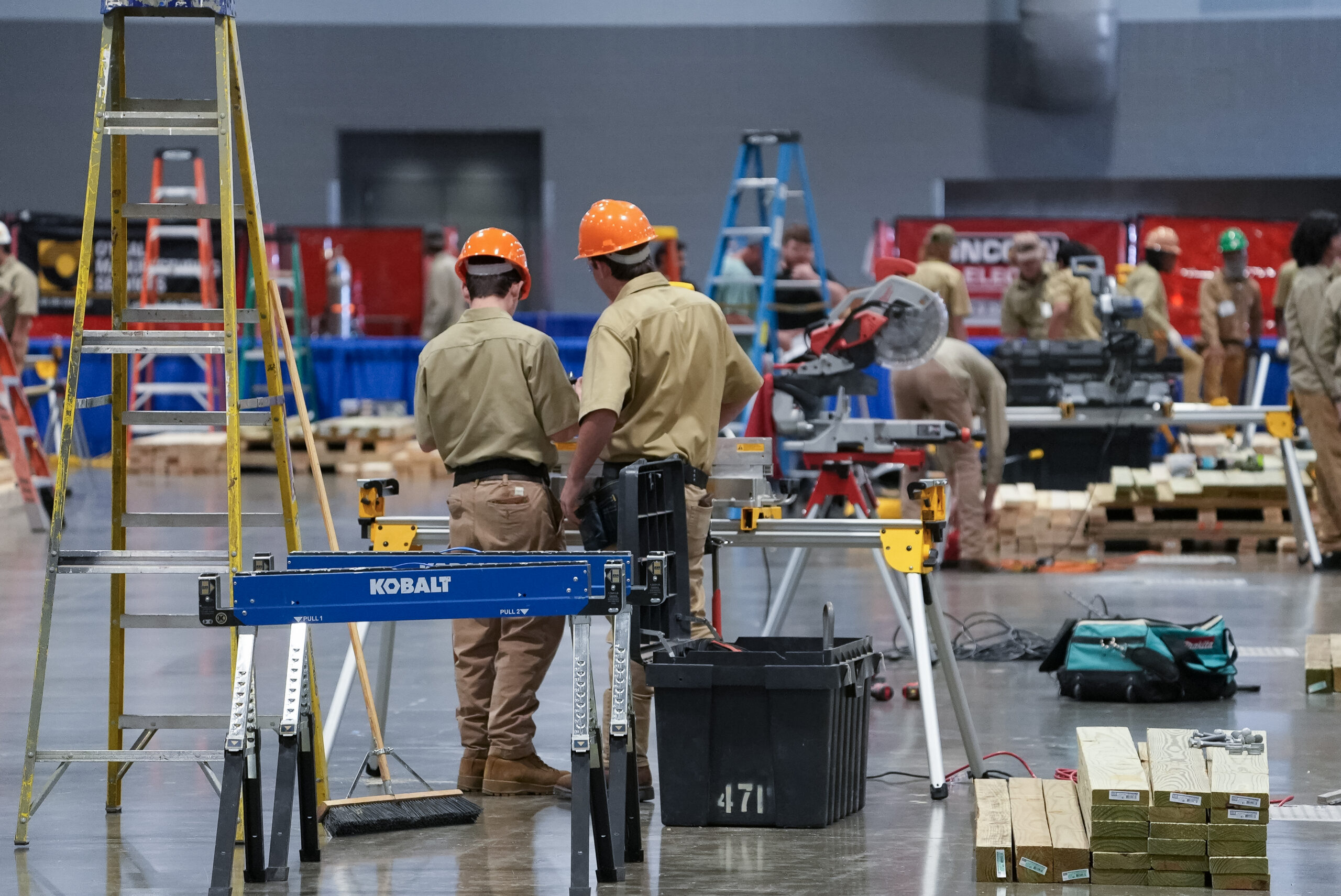 A teenage boy with curly hair wears safety glasses and smirks while using a wood planer to cut wood into the right size in what looks like a high school shop class.